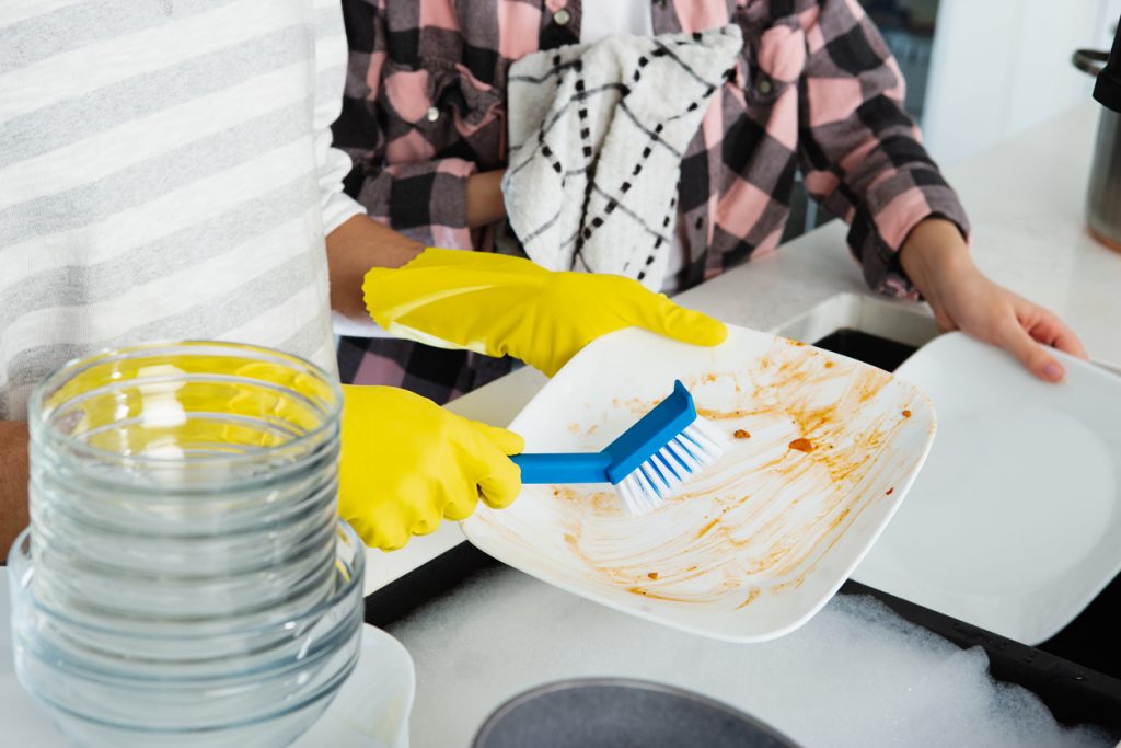 Two people in the kitchen washing dishes and removing food waste from plates.