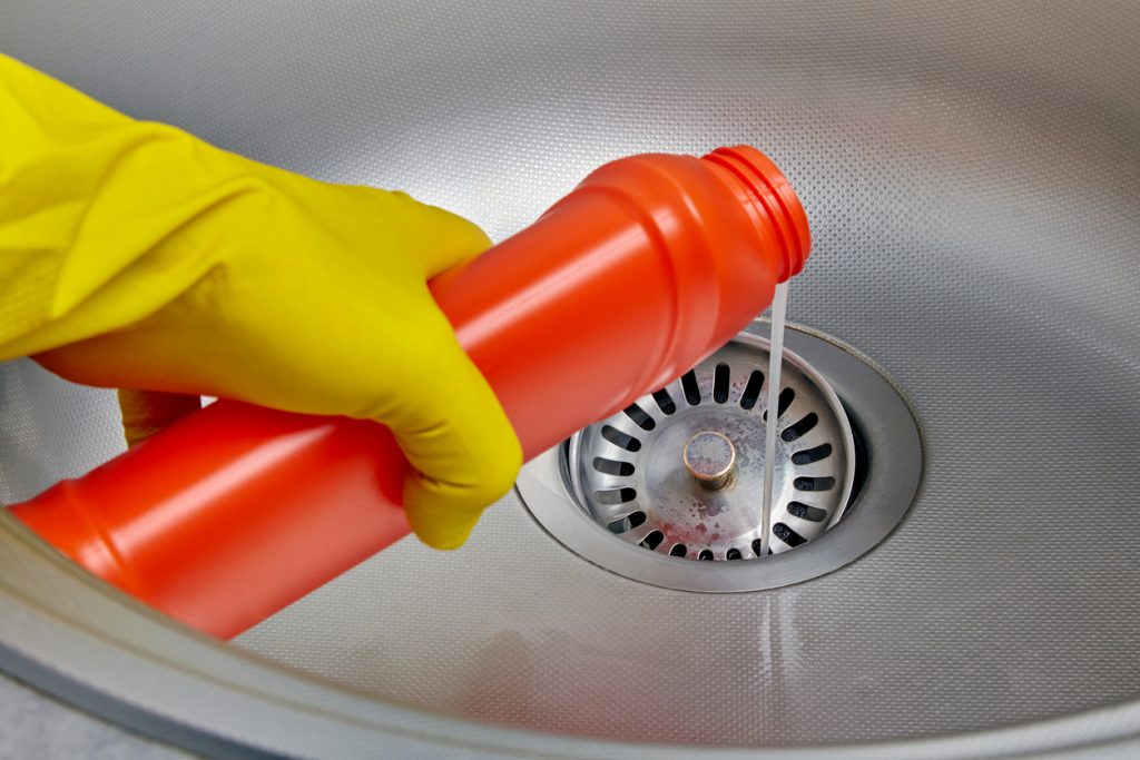 Preventing drain clogs - Person wearing a glove while pouring cleaner down the drain of a kitchen sink.